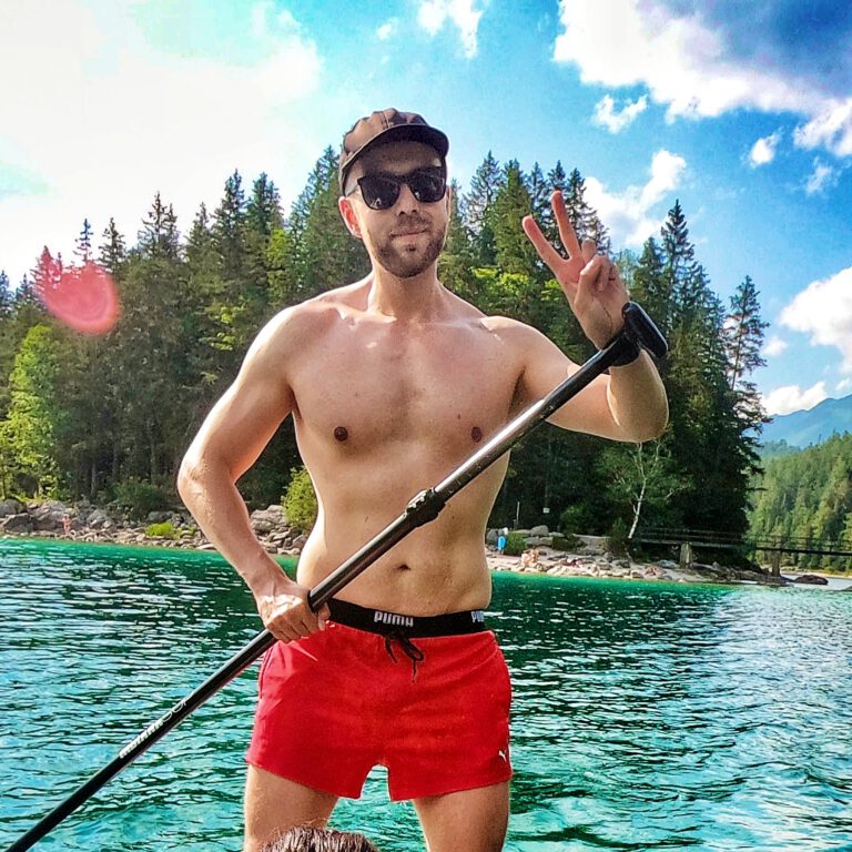 Mann mit schwartem Cap, Sonnenbrille und roter Badehose zeigt Peace-Zeichen, während er auf einem SUP steht.